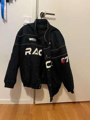 Svart racing vindjacka med tryck - Cool svart vindjacka med vit och röd racing-inspirerad text och detaljer. Jackan har hög krage, tryckknappar och ribbade muddar. Materialet är syntet och designen är sportig med kontrastlinjer och logotyp på bröstet och ärmen. Storlek M 🖤