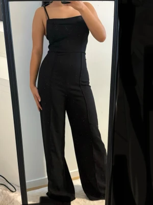 Svart jumpsuit med raka ben - Säljer en svart jumpsuit med smala axelband och raka, vida ben. Jumpsuiten har en enkel och stilren design med markerad midja och slät yta. Perfekt för dig som gillar minimalistiskt och trendigt mode.
