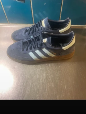 Adidas Originals navy sneakers - Mörkblå Adidas Originals sneakers i mocka med klassiska vita ränder på sidorna och vit hälflik. Skorna har brun gummisula och snörning framtill. Perfekta för dig som gillar retrostil och streetwear.