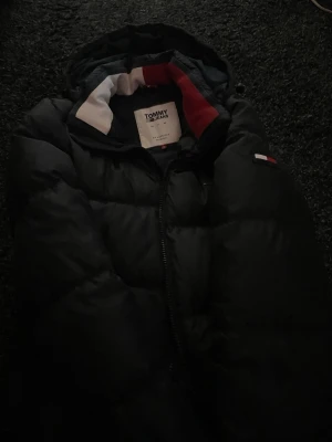 Svart dunjacka från Tommy Jeans - Snygg svart dunjacka från Tommy Jeans med huva och dragkedja. Jackan har klassiska färgdetaljer i rött, vitt och blått på insidan av kragen samt en liten logga på ärmen. Perfekt för kalla vinterdagar och riktigt stilren.