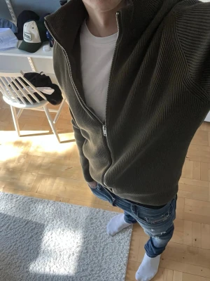 Olivgrön ribbad ziptröja - Säljer en olivgrön ribbad ziptröja med hög krage och hel dragkedja framtill. Tröjan har en clean look utan synliga loggor och är tillverkad i ett tjockare stickat material. Perfekt för lager på lager och enkel att matcha med jeans eller t-shirt.