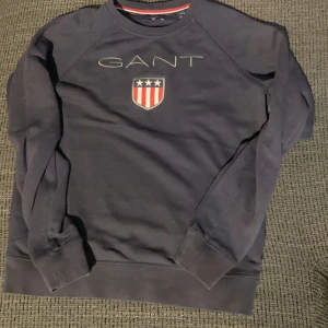 Mörkblå sweatshirt från Gant - Mörkblå sweatshirt från Gant med klassiskt tryck och sköld på bröstet. Tröjan har rund halsringning, långa ärmar och ribbade muddar. Perfekt för dig som gillar stilren look