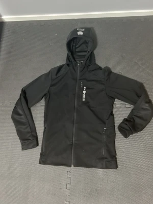  Sail Racing - Cool svart vindjacka från Sail Racing med huva och hel dragkedja. Jackan har en bröstficka med dragkedja och logotyp på framsidan. Materialet är slätt och funktionellt, perfekt för aktiva dagar utomhus. Storlek medium och den har en liten skada men syns knappt finn en bild på det