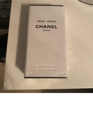 Chanel Paris-Venise Eau de Toilette - Chanel Paris-Venise Eau de Toilette i en elegant vit rektangulär kartong med svarta detaljer. Förpackningen har en lyxig och stilren design, typisk för Chanel. Volymen är 125 ml och parfymen är inspirerad av resan mellan Paris och Venedig.