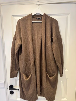 Brun lång kofta från 157 - Mysig lång kofta i brun nyans från 157. Koftan har öppen front, två stora fickor framtill och ribbade muddar vid ärmslut och nederkant. Perfekt för lager på lager och chill dagar. Materialet är mjukt och stickat för extra komfort.