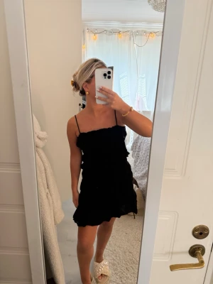 Svart kort klänning med volang - Säljer en svart kort klänning med tunna axelband och volangdetaljer. Klänningen har en feminin och enkel design som passar perfekt till sommaren. Materialet känns lätt och luftigt, vilket gör den skön att bära under varma dagar.