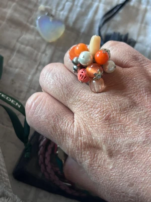 Färgglad ring med pärlor  - Unik ring med kluster av pärlor  i orange, vitt, silver och rosa. Ringen har en lekfull design med olika former och material, bland annat metall och plast. Perfekt för dig som gillar att sticka ut med accessoarer.
