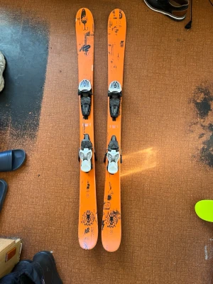 Orange skidor från K2 med bindningar - Säljer ett par orangea skidor från K2 med svarta och vita bindningar. Skidorna har coola svarta grafiska detaljer och är tillverkade i ett tåligt material som passar för alpina äventyr. Perfekt för dig som vill sticka ut i backen.