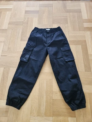 Svarta cargobyxor med resår - Snygga svarta cargobyxor med resår i midjan och vid bensluten. Byxorna har stora sidofickor med lock på båda benen och två bakfickor. Perfekta för en avslappnad streetwear-look. Tillverkade i ett slitstarkt bomullstyg.