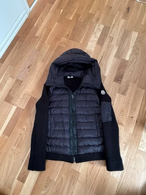 Moncler Cardigan - Snygg svart pufferjacka från Moncler med quiltad front och huva. Jackan har långa stickade ärmar, dragkedja framtill och en Moncler-logga på ärmen. Perfekt för kyliga dagar och riktigt stilren look.