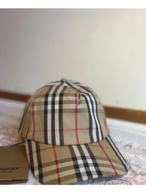 Beige rutig keps från Burberry - Säljer en klassisk keps från Burberry med ikoniskt beige, svart, vitt och rött rutmönster. Kepsen har böjd skärm och justerbar rem bak med Burberry London England-märke. Perfekt accessoar för dig som gillar stilrena och tidlösa detaljer.