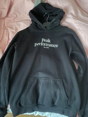 Svart hoodie från Peak Performance - Svart hoodie från Peak Performance med vit logga och text på bröstet. Klassisk känguruficka framtill och huva. Perfekt för en chill och sportig stil. Mjuk insida och ribbade muddar vid ärmslut och nederkant.