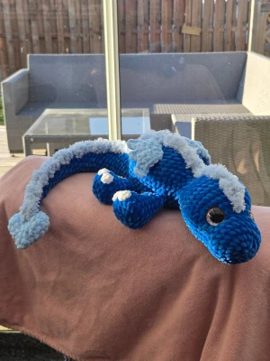 Blå virkad sagodrake - Supermjuk och gosig drake i blått och ljusblått sammetsgarn. Draken har stora glittriga säkerhetsögonögon, vita klor och en lång svans med ljusblå taggar längs ryggen. Perfekt som dekoration eller kramkompis. 