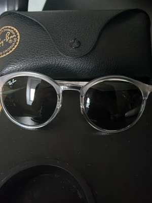  Ray-Ban solglasögon - Knappt använda inga repor Säljer ett par Ray-Ban solglasögon med rund form och transparent båge. Glasen är mörka och ger en clean look. Kommer med original Ray-Ban fodral. Perfekta för dig som vill ha en stilren och modern accessoar till sommaren.