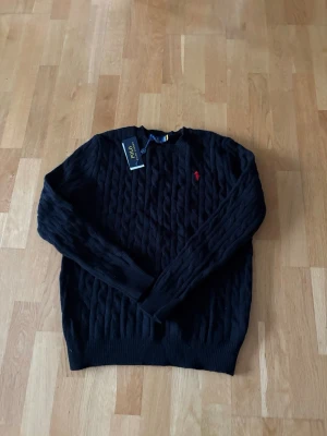 Svart kabelstickad tröja Polo Ralph Lauren - Kabelstickad svart tröja från Polo Ralph Lauren med rund halsringning och röd broderad logga på bröstet. Tröjan har långa ärmar och ribbade muddar vid ärmslut och nederkant. Perfekt för dig som gillar klassisk och stilren design.
