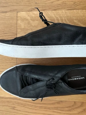 Svarta sneakers från Vagabond 40 - Stilrena svarta sneakers från Vagabond med vit sula och diskret ormmönstrad struktur i materialet. Skorna har klassisk låg profil, svarta snören och en modern look som passar till det mesta. Perfekta för dig som gillar minimalistisk design.