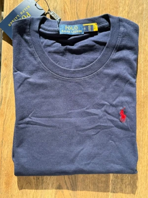 Marinblå t-shirt från Polo Ralph Lauren - Snygg marinblå t-shirt från Polo Ralph Lauren med klassisk rund halsringning och det ikoniska röda broderade logotypen på bröstet. Tillverkad i mjuk bomull för en skön känsla och perfekt passform. Enkel och stilren design som funkar till allt.