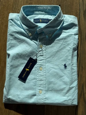 Ljusblå skjorta från Ralph Lauren - Säljer en klassisk ljusblå skjorta från Ralph Lauren med button-down krage och broderad logga på bröstet. Skjortan har långa ärmar, vita knappar och är tillverkad i mjuk bomull. Perfekt för en clean och stilren look.