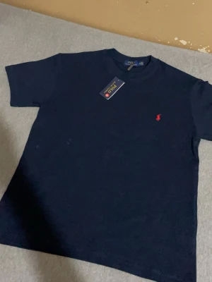 Marinblå t-shirt från Polo Ralph Lauren - Klassisk svart t-shirt från Polo Ralph Lauren med röd broderad logga på bröstet. T-shirten har rund halsringning och korta ärmar. Tillverkad i mjuk bomull för en skön känsla och enkel stil.