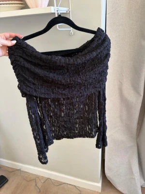 Svart offshoulder topp i spets - Cool svart offshoulder topp med transparent spets och struktur. Toppen har långa ärmar och ett brett, draperat axelparti som ger en unik look. I storlek S från Nelly💕
