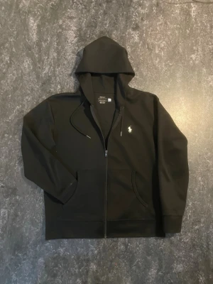 Svart hoodie från Polo Ralph Lauren - (Priset kan diskuteras) Svart hoodie från Polo Ralph Lauren med klassisk logga broderad på bröstet. Hoodien har dragkedja, fickor framtill och justerbar huva med snörning. Perfekt för en avslappnad och cool look.