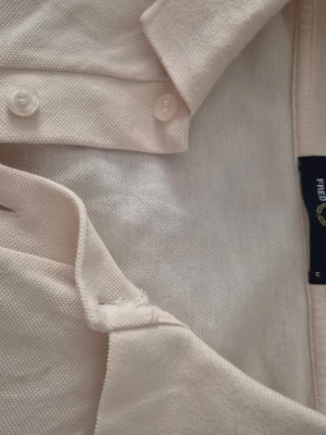 Rosa pikétröja från Fred Perry - Klassisk rosa pikétröja från Fred Perry med kort ärm, krage och knappar framtill. Tröjan har broderad logga på bröstet och är gjord i bomull med pikéstruktur. Perfekt för en clean och sportig stil.