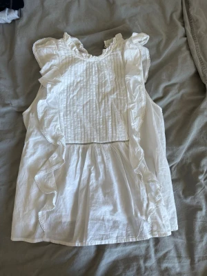 Vit ärmlös blus med volanger - Supersöt vit blus med volanger längs sidorna och runt halsen. Säljer pga att den blivit för liten. Blusen kommer från Bikbok. 💗