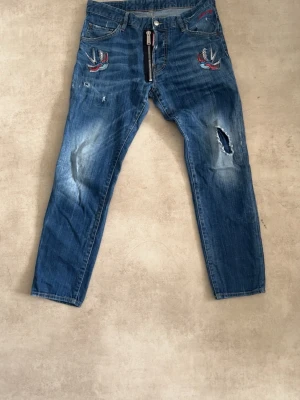 Dsquared2 jeans - Säljer ett par blå jeans med slitningar och hål, samt coola broderade svalor på framsidan. Jeansen har dragkedja framtill och klassisk femficksmodell. Materialet är denim i bomull och passformen är rak.