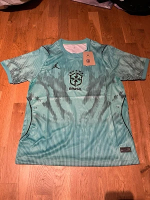 Turkos Brasil tränings t-shirt Jordan - Snygg turkos tränings t-shirt från Jordan i samarbete med Brasil. Tröjan har svart grafiskt mönster, rund halsringning och korta ärmar. Brasil-logga och Jordan-logga på bröstet. Tillverkad i lätt och ventilerande material, perfekt för träning eller match.