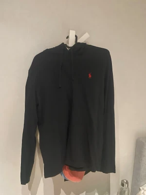 Svart hoodie från Ralph Lauren - Klassisk svart hoodie från Ralph Lauren med röd broderad logga på bröstet. Hoodien har långa ärmar, huva med snörning och är tillverkad i mjuk bomull. Perfekt för en avslappnad och stilren look. Denna hoodie är lite tunnare vilket gör det skönare att ha den på sommarkvällar