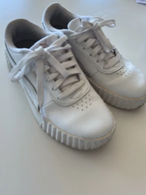 Vita sneakers från Puma - Säljer ett par vita sneakers från Puma med klassisk design och ribbad sula. Skorna har snörning, perforerade detaljer på tån och är tillverkade i läder. Perfekta för dig som gillar stilrena och sportiga skor.