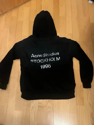 Svart hoodie Acne Studios - Säljer en svart hoodie från Acne Studios med tryck på ryggen där det står 'Acne Studios STOCKHOLM 1996'. Klassisk passform med huva och ribbade muddar. Perfekt för dig som gillar stilrena och trendiga plagg. Materialet känns mjukt och bekvämt.