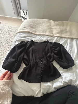 Svart blus med puffärmar - Säljer en svart blus med v-ringning och knappar framtill. Blusen har puffiga långärmar och en figursydd midja som ger en snygg siluett. Materialet är lätt och strukturerat, perfekt för att skapa en cool look.