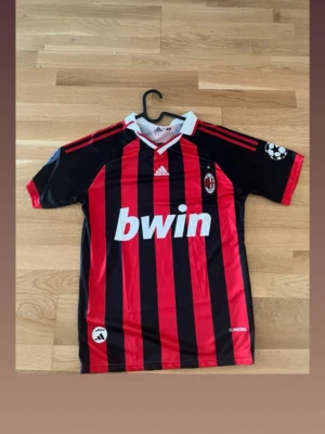 AC Milan Ronaldinho matchtröja - Säljer en klassisk AC Milan matchtröja från Adidas med Ronaldinho och nummer 80 på ryggen. Tröjan har röda och svarta vertikala ränder, vit krage och Champions League-märke på ärmen. Materialet är lätt och andas, perfekt för fotboll eller samling.