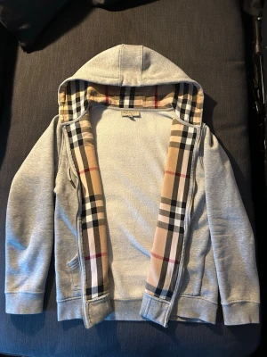 Grå hoodie från Burberry med dragkedja - Säljer en grå hoodie från Burberry i herr storlek M/S med klassiskt rutigt foder i luvan och längs dragkedjan. Hoodien har broderad logga på bröstet, ribbade muddar och två fickor. Materialet är mjukt och bekvämt. 