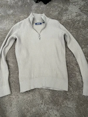 Ljusgrå stickad tröja med dragkedja - Säljer en ljusgrå stickad tröja från Jack & Jones med ribbad struktur och halv dragkedja framtill. Tröjan har hög krage och långa ärmar, perfekt för att hålla värmen. Materialet är mjukt och stretchigt, vilket gör den bekväm att bära.