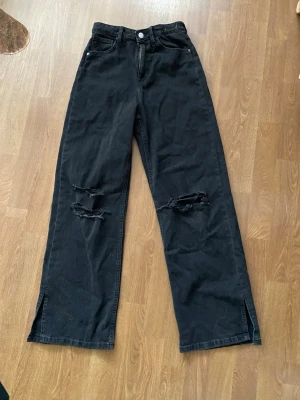 Svarta wide leg jeans med slitningar - Coola svarta jeans med wide leg passform och slitningar på knäna. Jeansen har hög midja, klassiska fickor och slitsar längst ner på benen. Perfekta för dig som gillar en avslappnad och trendig look.