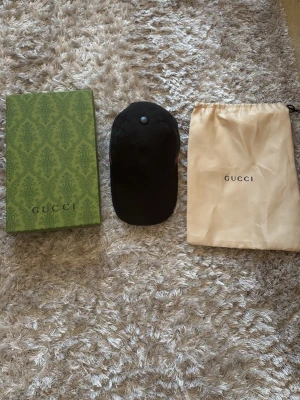 Svart keps från Gucci med logga - Säljer en svart keps från Gucci med diskret GG-monogram över hela kepsen. Klassisk böjd skärm och justerbar rem bak. Sidorna har Guccis ikoniska gröna och röda ränder. Kepsen kommer med originalkartong och dustbag.
