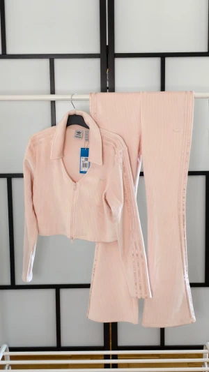 Velour ribbad tränings set Adidas  - Superfint och lyxigt Adidas‑set i mjuk, ribbad velour. Både jacka och byxor har klassiska tre ränderna längs sidorna och broderad logga. Byxorna har lätt flare för en smickrande siluett och jackan är en zip‑modell med krage.  Storlek: L   Skick: Helt nytt, lappar kvar   Färg: Ljusrosa    Perfekt när du vill gå från vardag till utekväll med noll ansträngning – sportigt, chic och lyxigt i ett och samma set. Nypris 1788 kr Mitt pris 1265 kr
