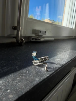Silvrig ormring med blå detaljer - Unik ring i form av en orm som slingrar sig runt fingret. Ringen är silvrig med detaljerade fjäll och har blå stenar på huvudet samt svarta ögon. Perfekt för dig som vill sticka ut med ett djurinspirerat smycke.