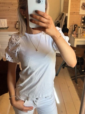 Vit topp med broderade puffärmar - Snygg vit topp med korta puffärmar i broderad spets och rund halsringning. Ärmarna har fina håldetaljer som ger en romantisk vibe. Perfekt att styla med jeans eller kjol för en fräsch look.