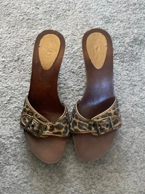 Leopardmönstrade träklack sandaler - Säljer ett par coola sandaler med träklack och leopardmönstrad rem med snyggt spänne. Remmen är i syntetmaterial och klacken är i mörkt trä.