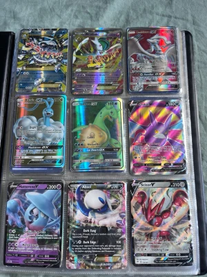 Pokémon GX och V samlarkort i pärm - Säljer en pärm med flera holografiska Pokémon GX och V samlarkort. Kort från olika generationer och med färgstarka motiv, bland annat Scizor, Absol, Hatterene, Grobat och fler. Perfekt för dig som vill utöka din samling med unika och glänsande kort.