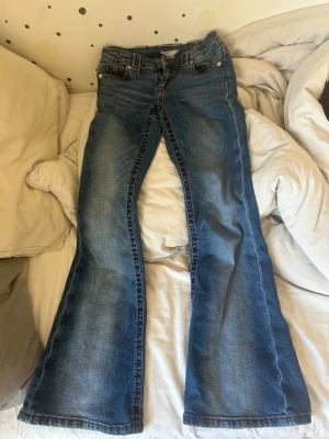 Blå bootcut jeans från Gina Tricot - Säljer ett par blå bootcut jeans från Gina Tricot med röda kontrastsömmar. Jeansen har klassisk femficksdesign, dragkedja och knapp framtill. Materialet är denim och passformen är utsvängd nedtill. Perfekt för dig som gillar retrostil!
