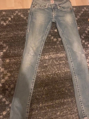 True Religion ljusblå jeans - Säljer ett par ljusblå jeans från True Religion med slitningar på benen och klassiska vita kontrastsömmar. Jeansen har raka ben och snygga broderade bakfickor med märkets logga på etiketten. Perfekt för dig som gillar streetstyle.