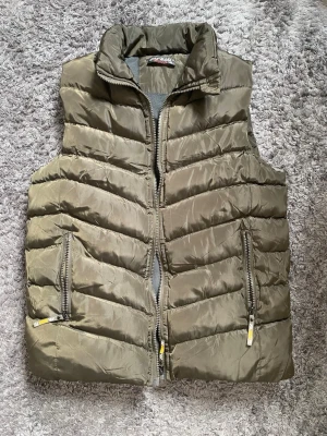 Olivgrön dunväst med dragkedja - Snygg olivgrön dunväst från Cacharel med hög krage och två fickor med dragkedja. Västen har ett quiltat mönster i chevron-stil och är tillverkad i glansigt material. Perfekt för lager-på-lager och håller dig varm under kyliga dagar.