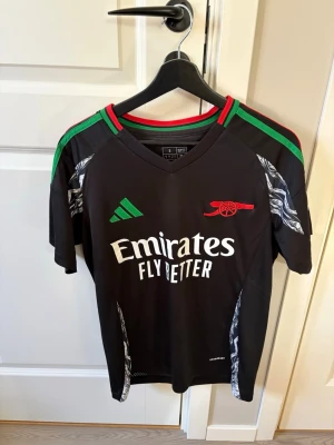 Arsenal svart matchtröja Adidas - Snygg svart Arsenal matchtröja från Adidas med gröna och röda detaljer på axlarna. Tröjan har klubbens logga och Emirates Fly Better-tryck på bröstet samt mönstrade inslag i vitt och grått på sidorna. Tillverkad i lätt och ventilerande material.