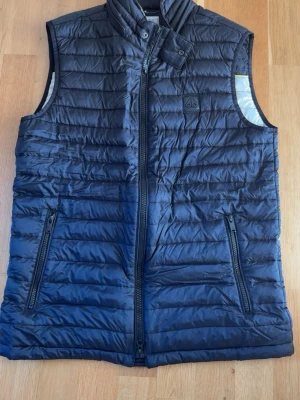 Svart dunväst från moose knuckles - Snygg svart dunväst från moose knuckles med quiltad design och hög krage. Västen har två praktiska fickor med dragkedja och diskret logotyp på bröstet. Perfekt för lager-på-lager och håller dig varm under kyliga dagar.