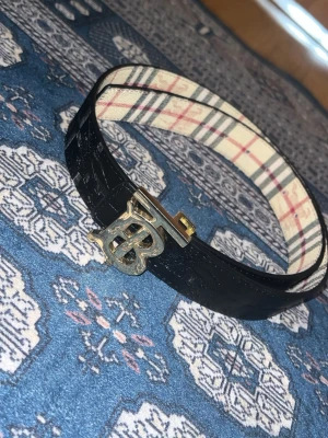 Burberry bälte med klassiskt mönster - Säljer ett snyggt Burberry bälte med ikoniskt rutigt mönster på ena sidan och svart skinn på andra. Spännet är i guld med Burberrys B-logo. Perfekt för att lyfta din outfit med en lyxig touch.