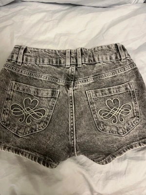 Grå denimshorts med broderade hjärtan - Säljer ett par gråa denimshorts med snyggt tvättad look. Baksidan har två fickor med broderade hjärtan och dekorativa sömmar. Säljer likadana blå fast annan storlek!
